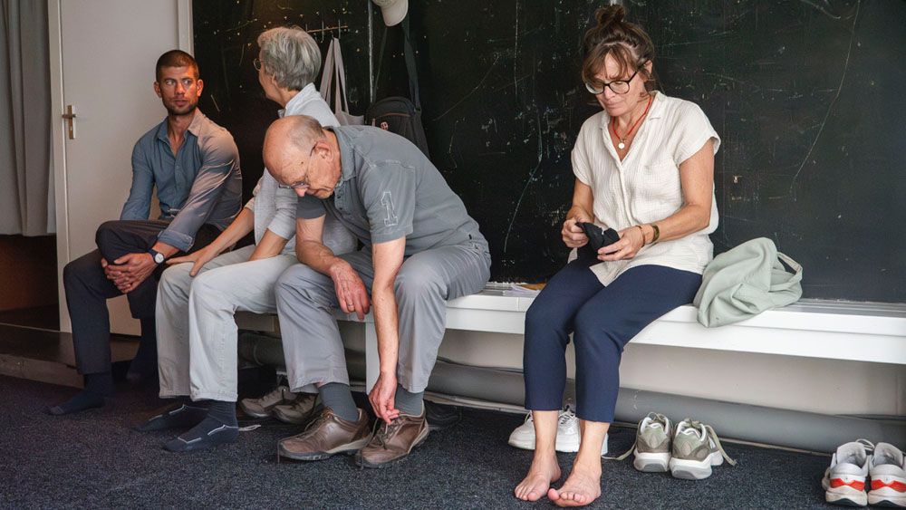 een groepje mensen zit op een bankje in de gang, ze trekken hun schoenen aan
